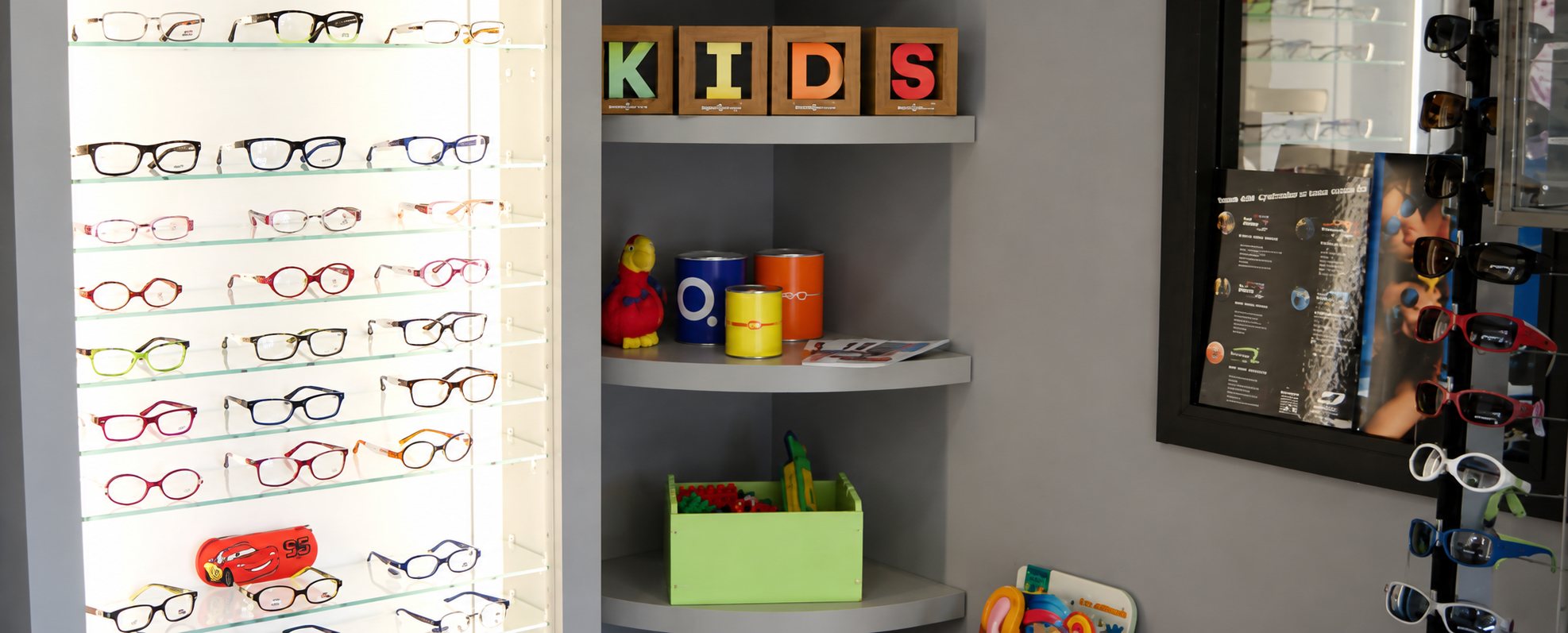 Coin enfants d'un magasin d'optique avec une vitrine lumineuse de lunettes, des cubes formant le mot KIDS et des étagères d'accessoires colorés.