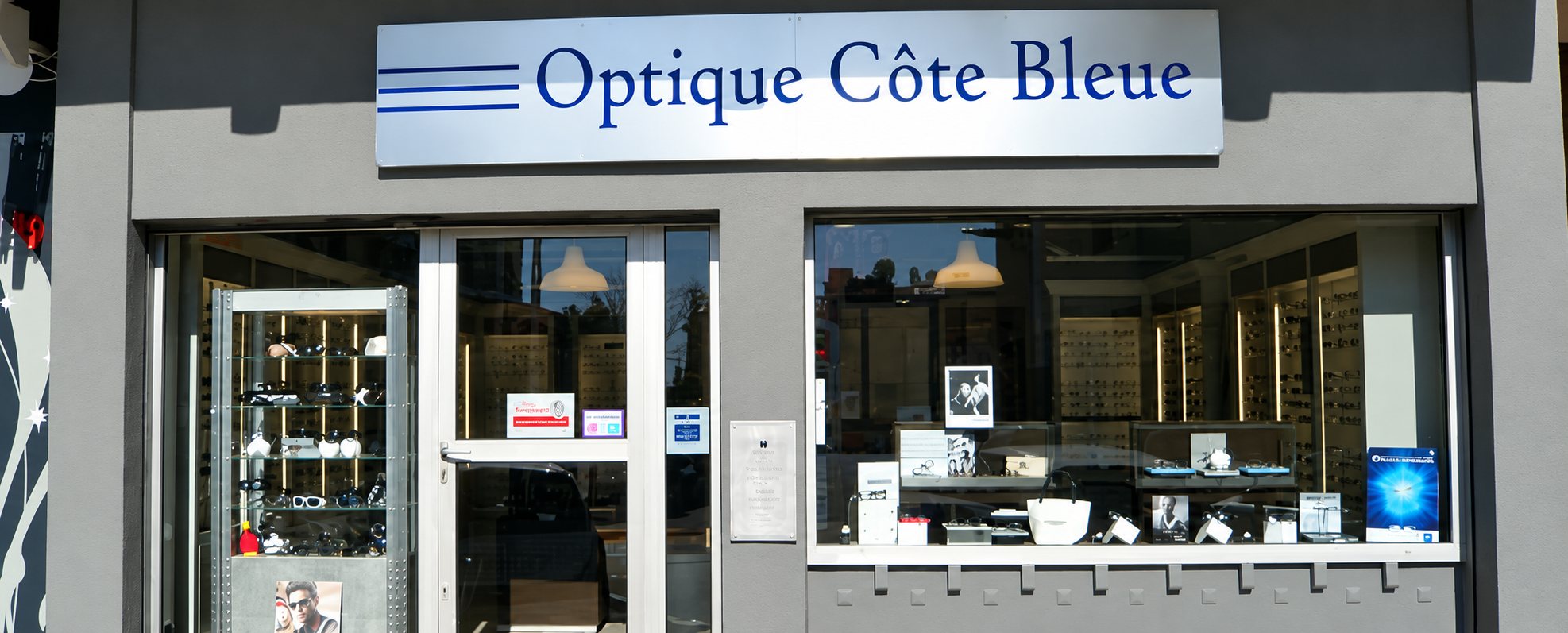 Façade grise du magasin Optique Côte Bleue, avec son enseigne argentée, sa porte vitrée et une vitrine présentant des lunettes.