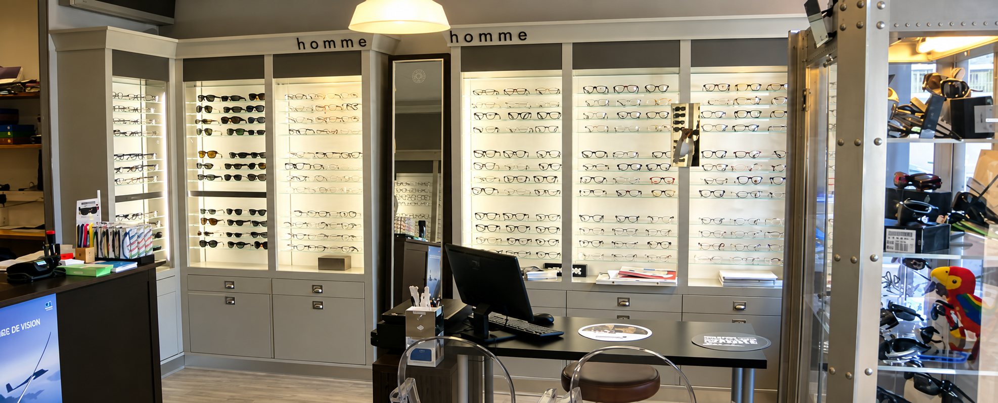 Intérieur d'une boutique d'optique avec des vitrines lumineuses remplies de lunettes de vue et de soleil, un bureau avec ordinateur et un comptoir au premier plan.