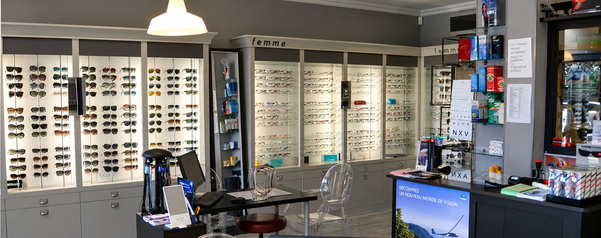 Intérieur de la boutique d’optique : à gauche un mur de lunettes de soleil, au centre les vitrines de montures de vue « femme », puis un bureau noir avec une chaise transparente et, à droite, le comptoir et ses présentoirs de produits.