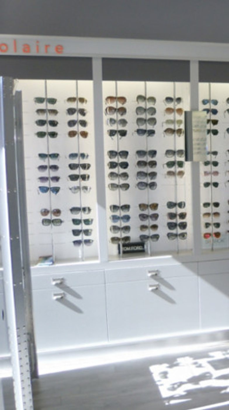 Un mur blanc d'une boutique d'optique présente de nombreuses paires de lunettes de soleil sur des supports, au-dessus de meubles de rangement blancs.